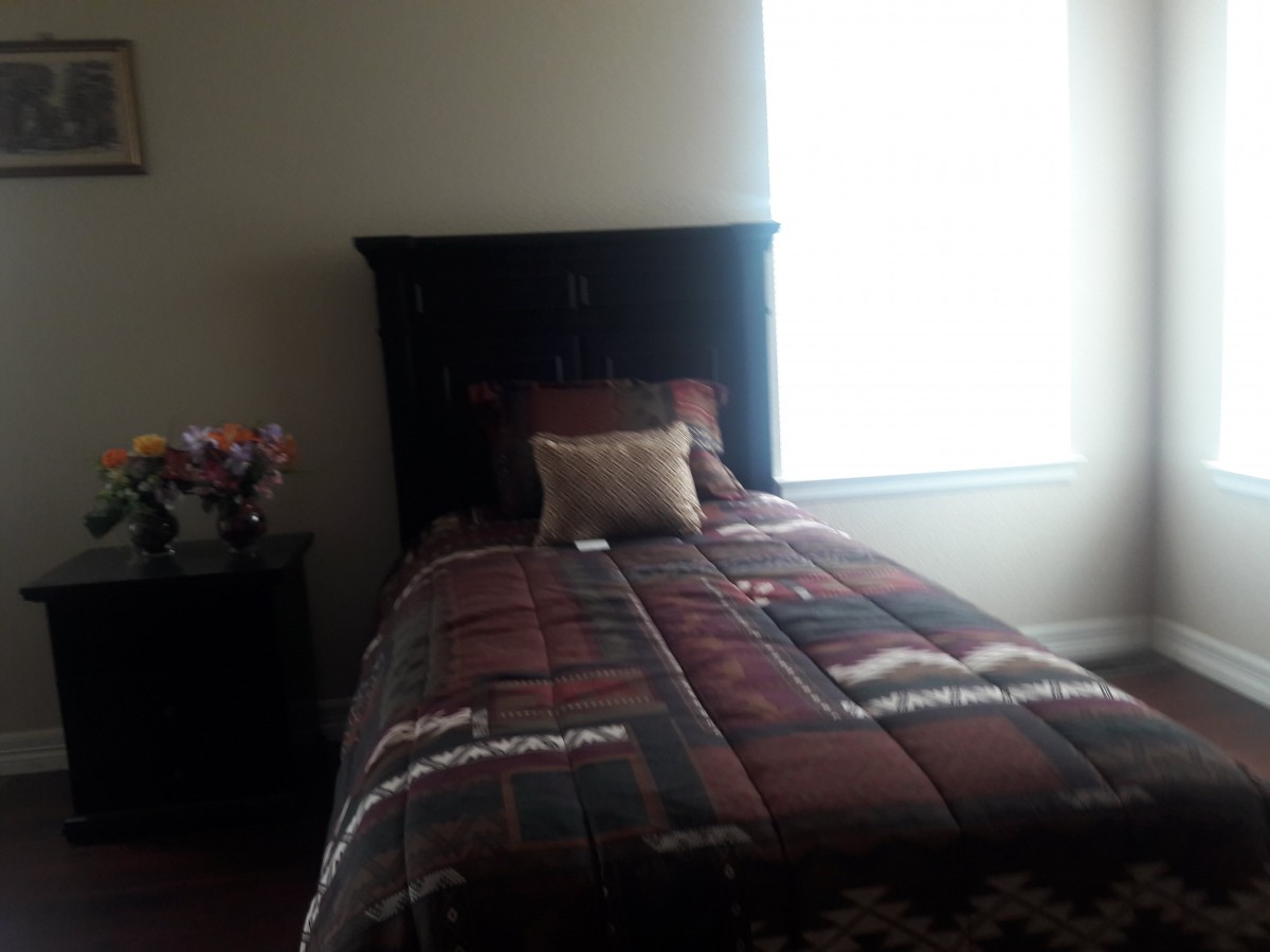 A cozy bedroom with a single bed, plaid comforter, and soft pillows next to a nightstand with flowers.