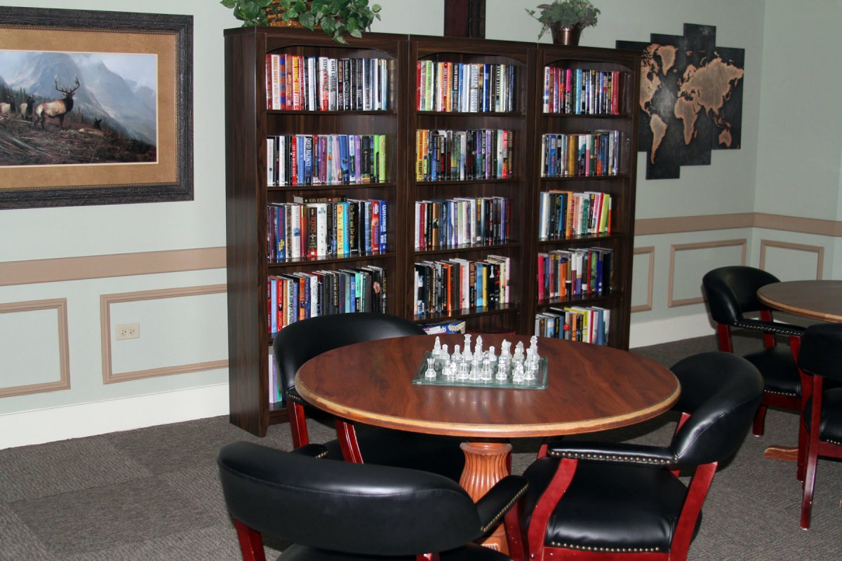 A cozy library corner featuring a round table with a chessboard, surrounded by bookshelves and chairs.