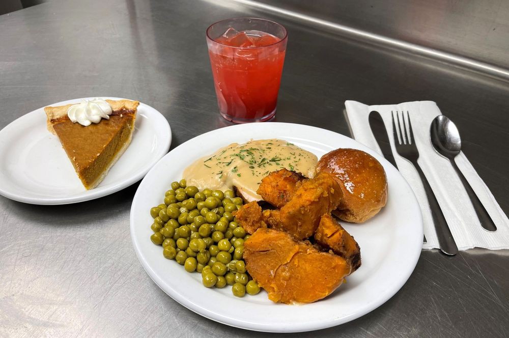 A dinner plate with turkey, gravy, peas, sweet potatoes, a roll, and a slice of pie served with a drink.