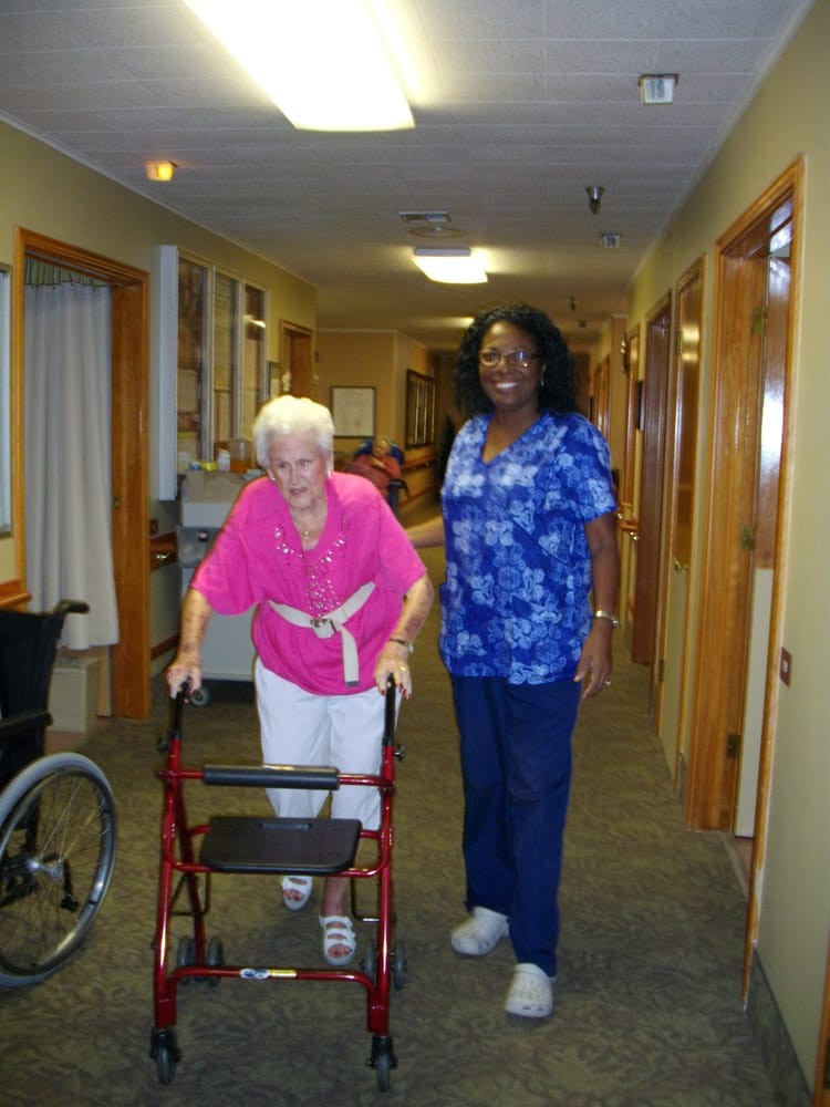 An elderly woman using a walker is accompanied by a nurse in a hallway of a care facility.