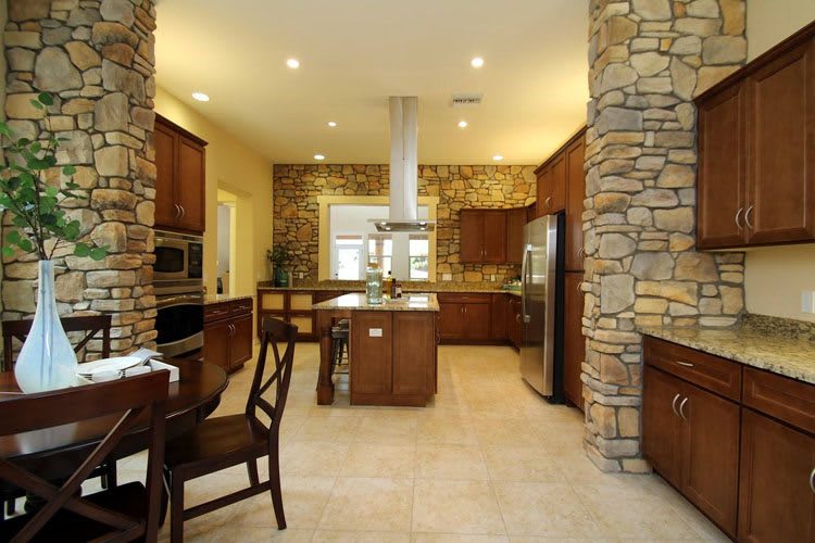 A spacious kitchen featuring stone walls, wooden cabinets, and an island counter, with a dining area and tiled flooring.