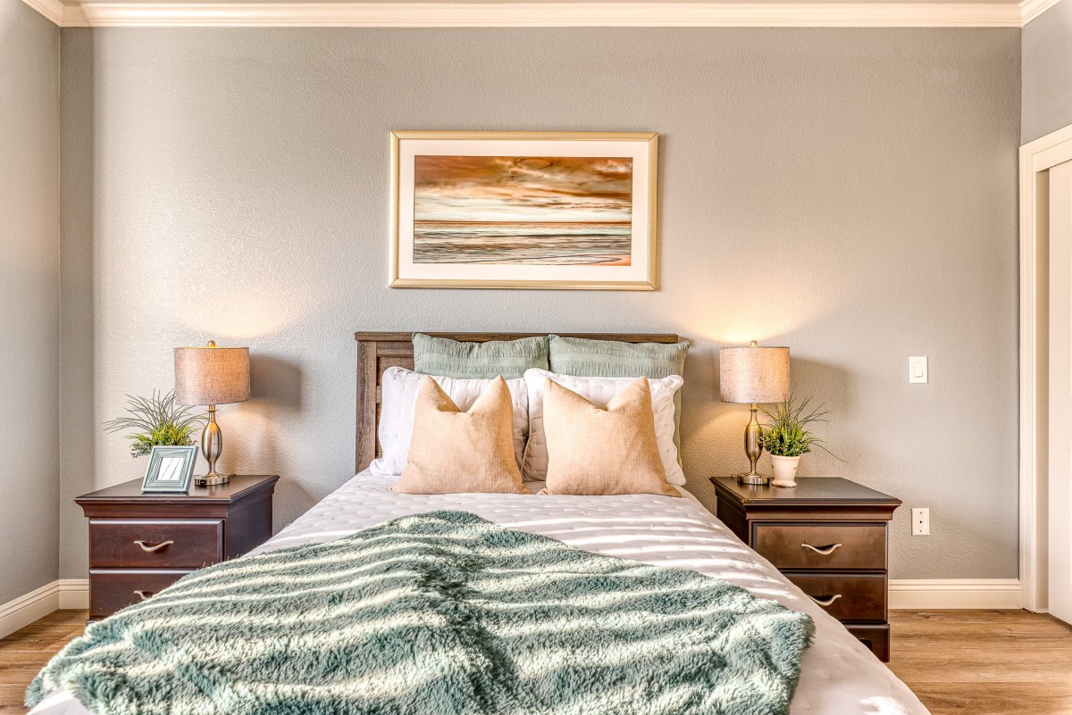 A cozy bedroom with a neatly made bed, flanked by two nightstands and lamps, featuring a landscape painting above the headboard.