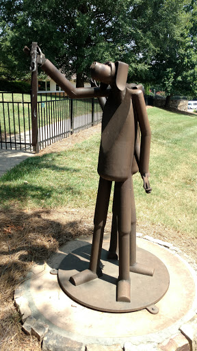 A metal abstract sculpture depicting a figure with one arm raised, set in an outdoor park.
