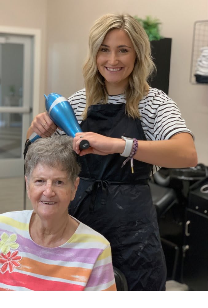 A hairstylist blow-drying a customer's hair while both are smiling in a salon.