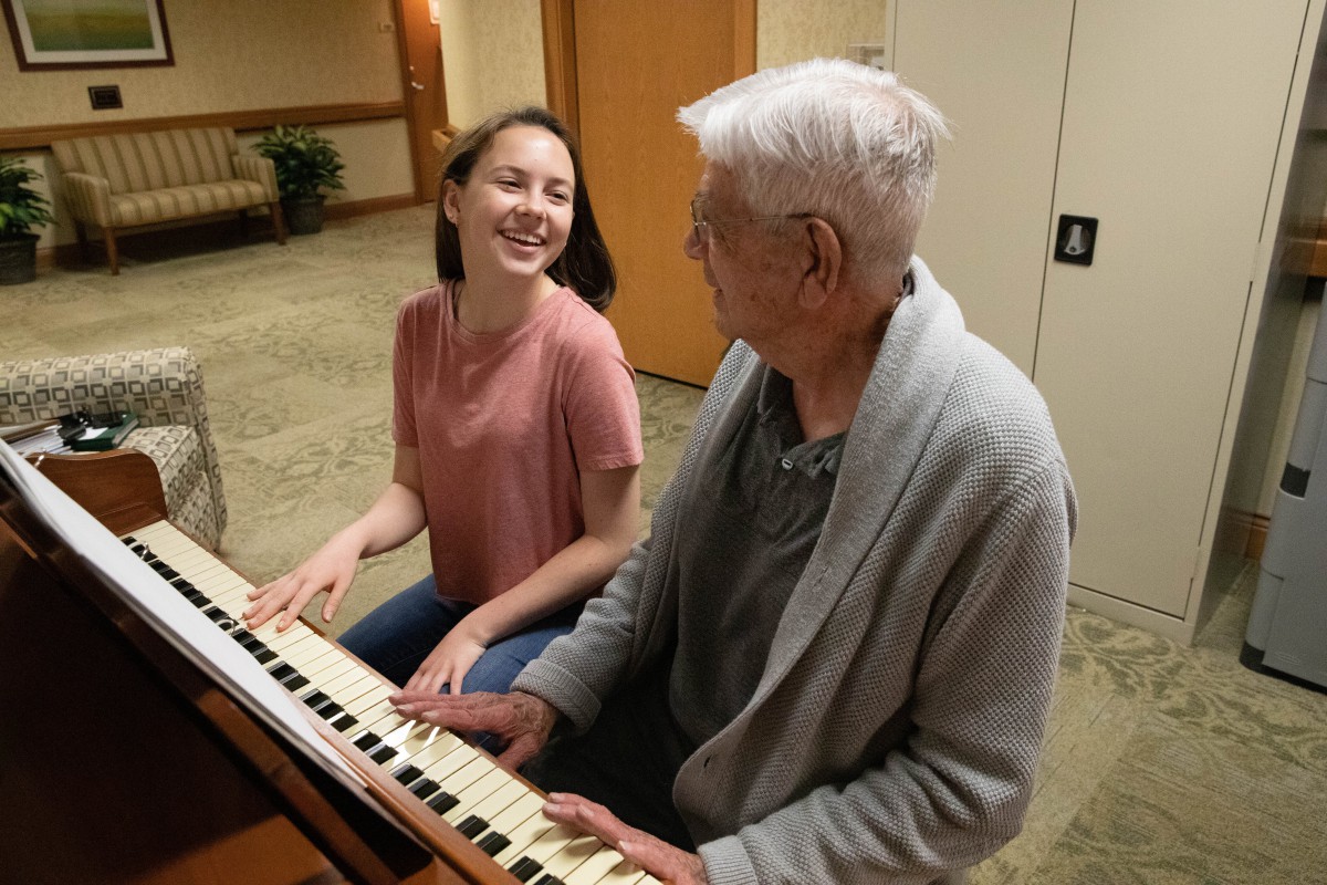 A young woman and an elderly man share a joyful moment playing the piano together indoors.