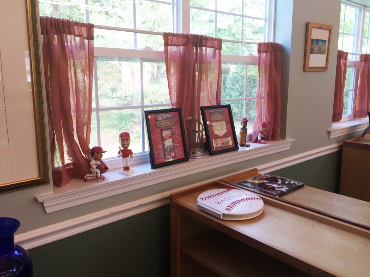 A room corner featuring a window with burgundy curtains, small decorative figures, framed photos on the windowsill, and a wooden desk with embroidery hoops.