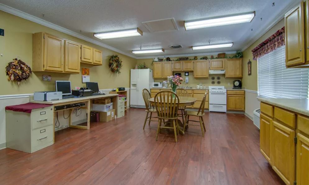 A multi-purpose room featuring a kitchen and office area with computers, a central wooden table, and chairs.