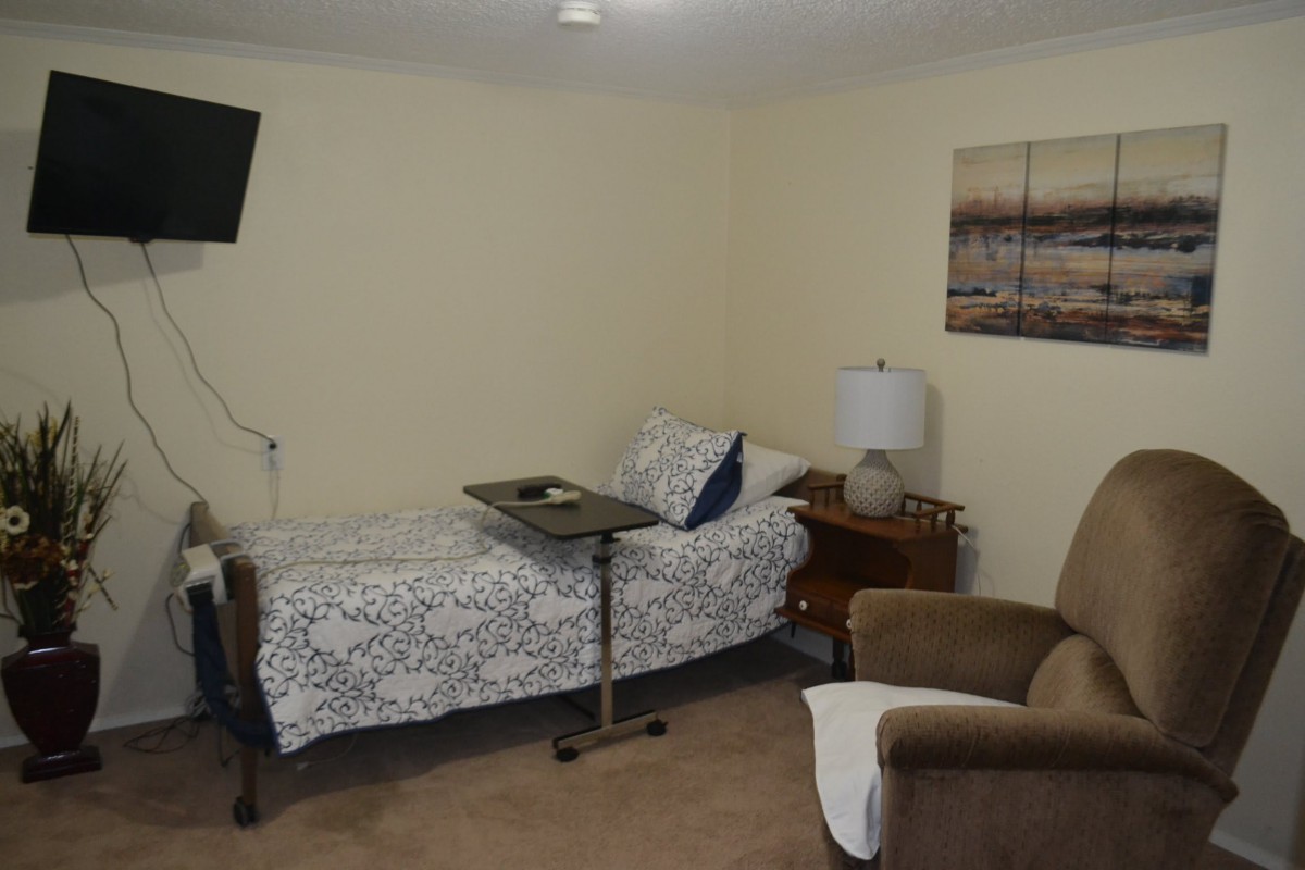 A cozy bedroom with a single bed, side table, armchair, and a television on the wall.