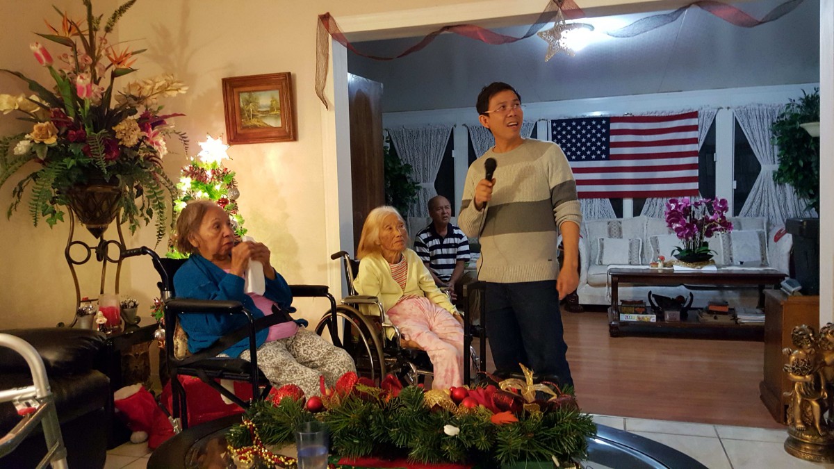 A family gathering with Christmas decorations and an American flag in the background, featuring a person speaking into a microphone.