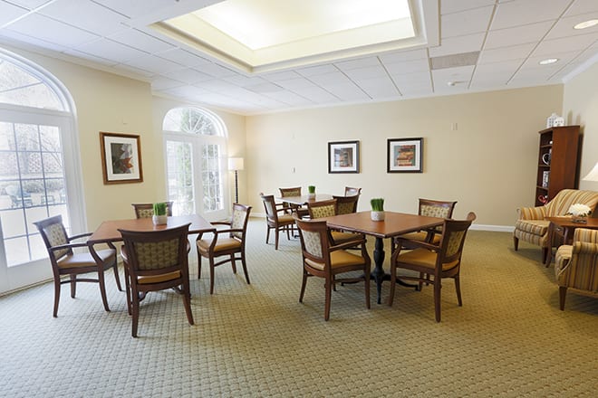 A cozy dining room with several tables and chairs arranged on a carpeted floor, illuminated by natural light through large windows.
