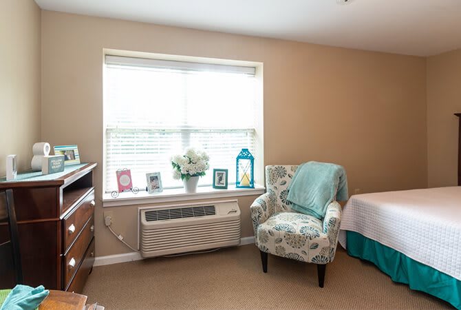 A cozy bedroom with a patterned armchair, large window, and neatly arranged decor.