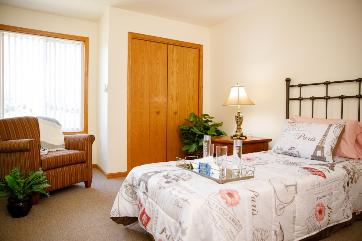 A cozy bedroom with a neatly made bed, bedside lamp, and comfortable chair near a window.