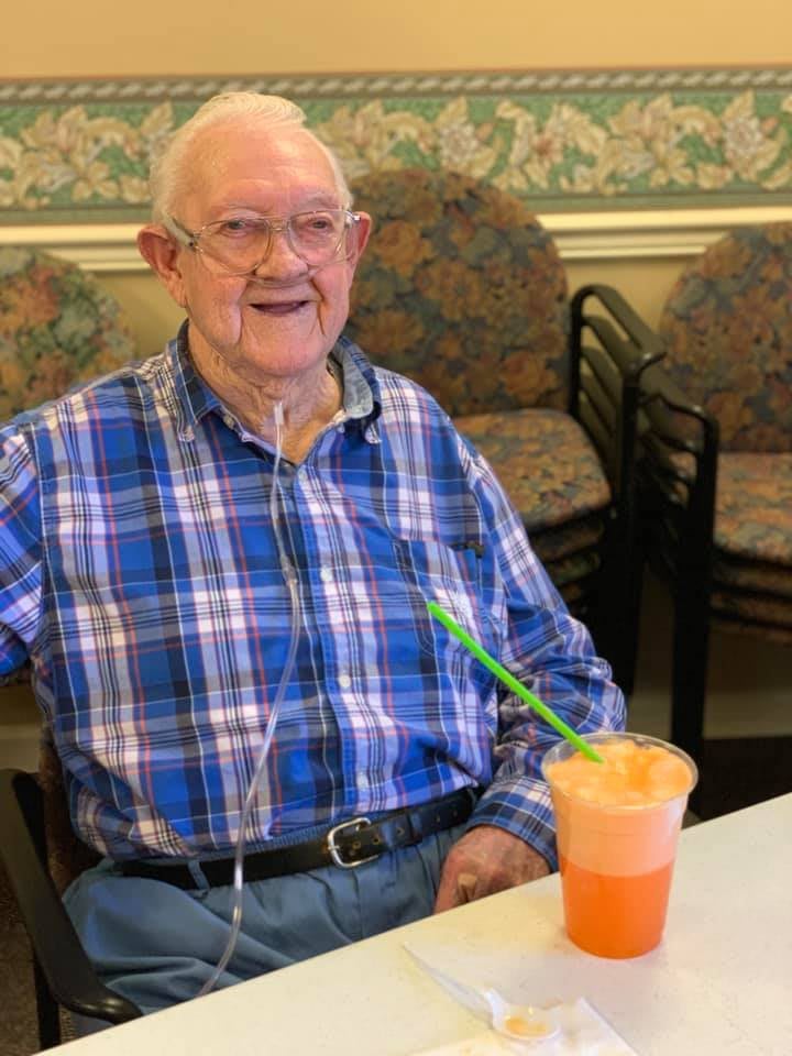An elderly man wearing a plaid shirt and glasses smiles while sitting at a table with a colorful drink.