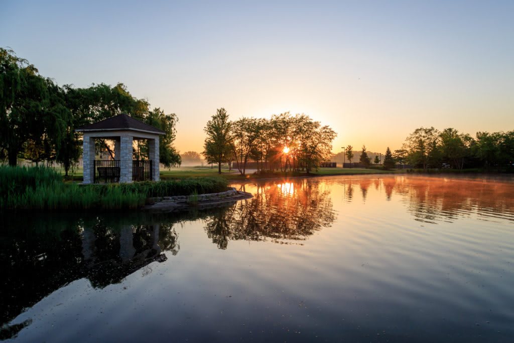 A serene sunset over a tranquil lake with a gazebo and trees reflected in the water.