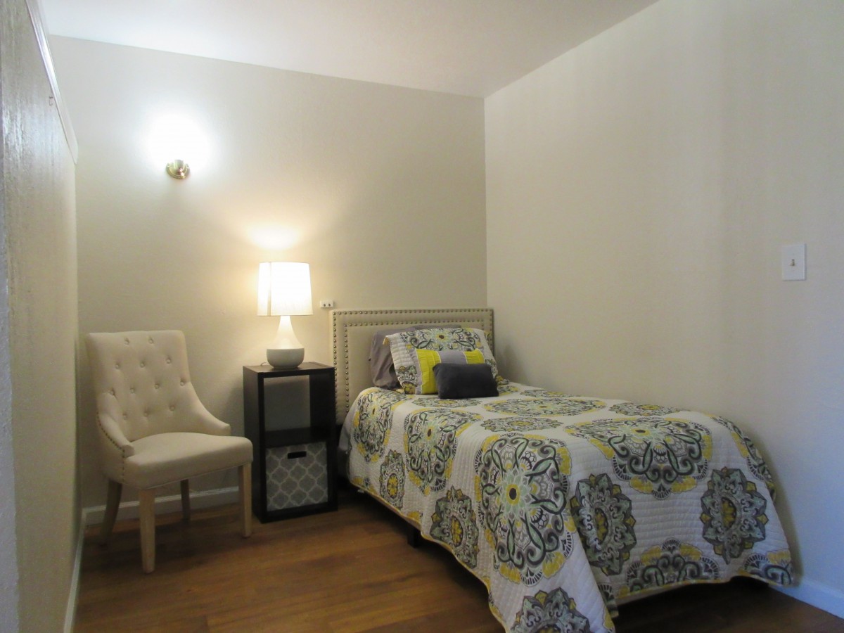 A cozy bedroom featuring a single bed with decorative bedding, a lamp on a nightstand, and an upholstered chair.