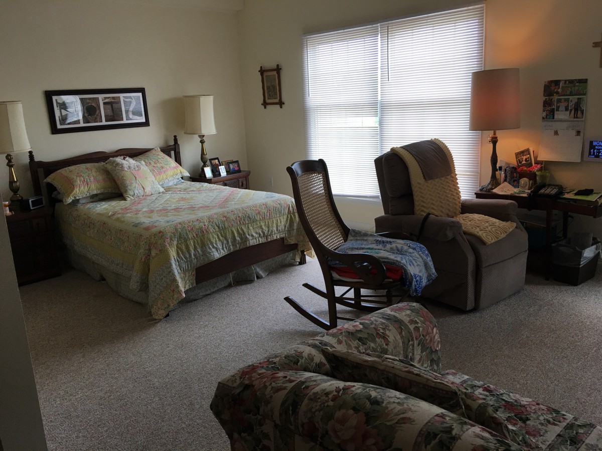 A cozy bedroom featuring a bed, rocking chair, armchair, and a lamp by the window.