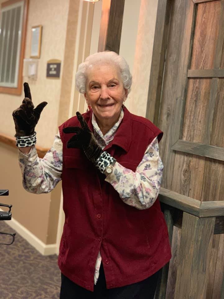 An elderly person smiling indoors, wearing a maroon vest and gloves.