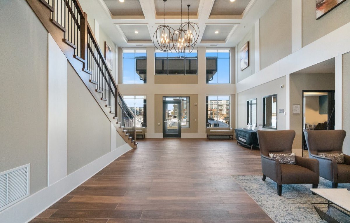 A spacious living room with high ceilings, a chandelier, hardwood floors, and large windows, featuring a staircase leading to the upper level.