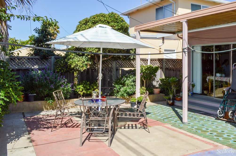 A sunny backyard patio with an umbrella-covered table and chairs, surrounded by a garden.