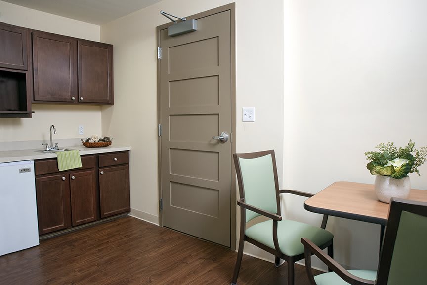 A small room with a kitchenette, featuring a wooden door, a table, and chairs.