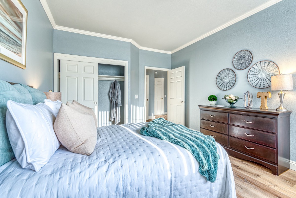 A cozy bedroom with blue walls featuring a neatly made bed, dresser, and decorative wall hangings.