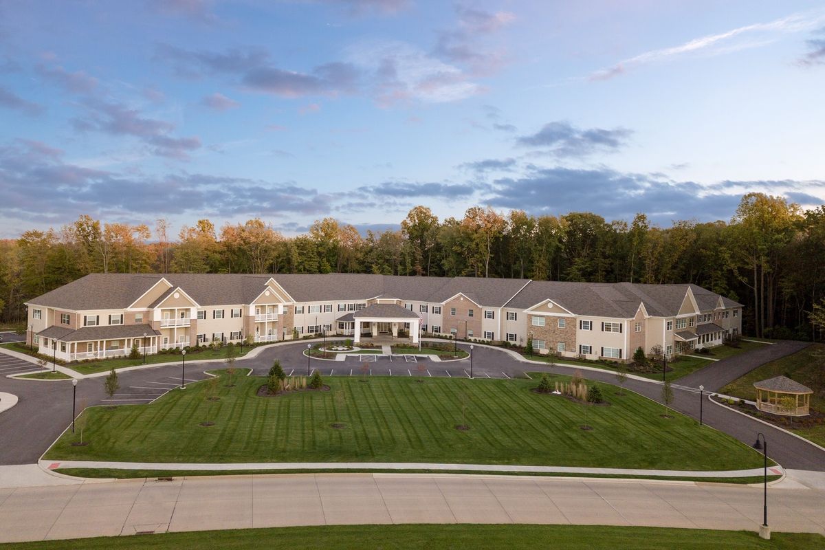 A large residential building surrounded by a manicured lawn and driveway, set against a backdrop of trees and a blue sky.