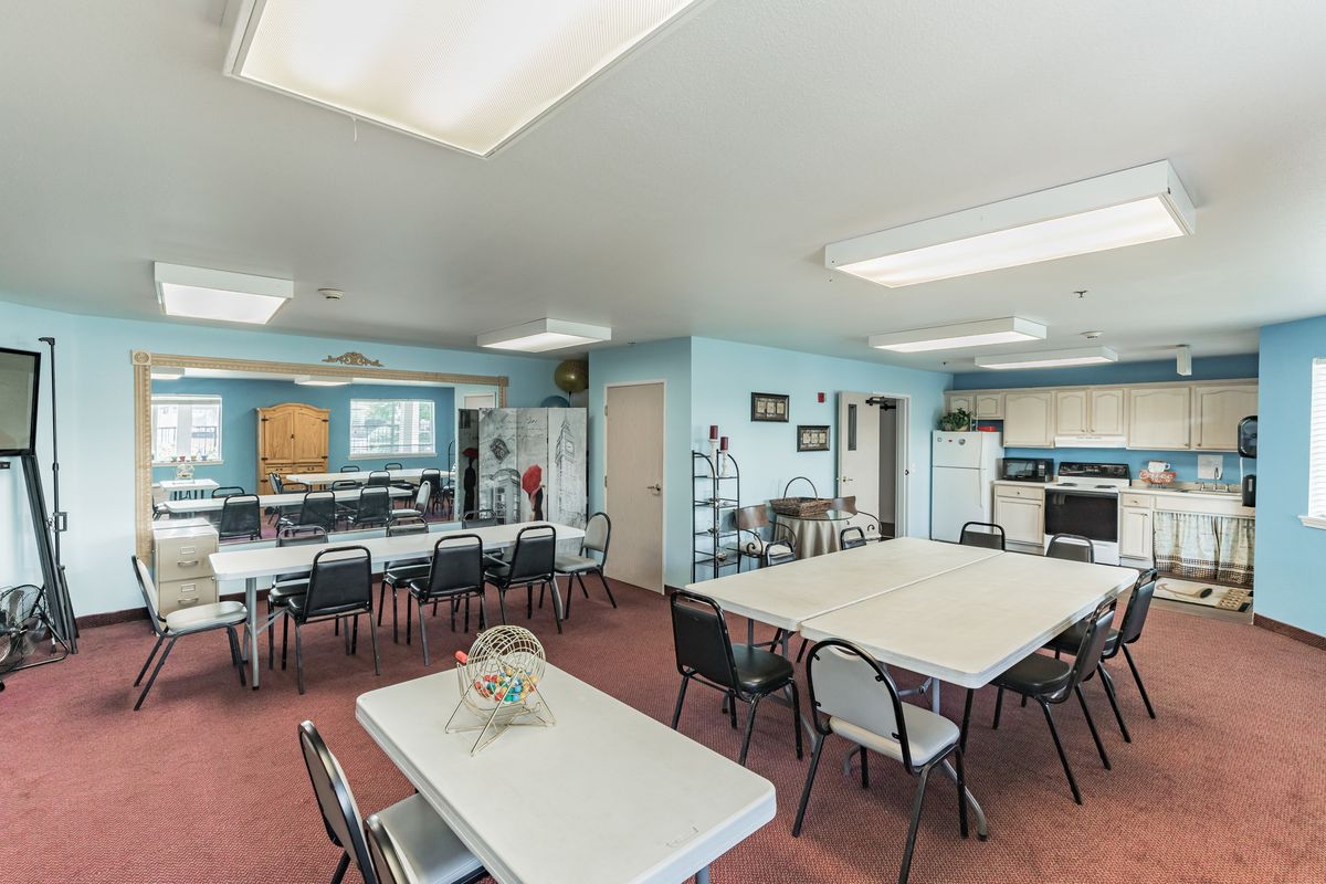 A community room featuring tables, chairs, and a small kitchen area with bright lighting.
