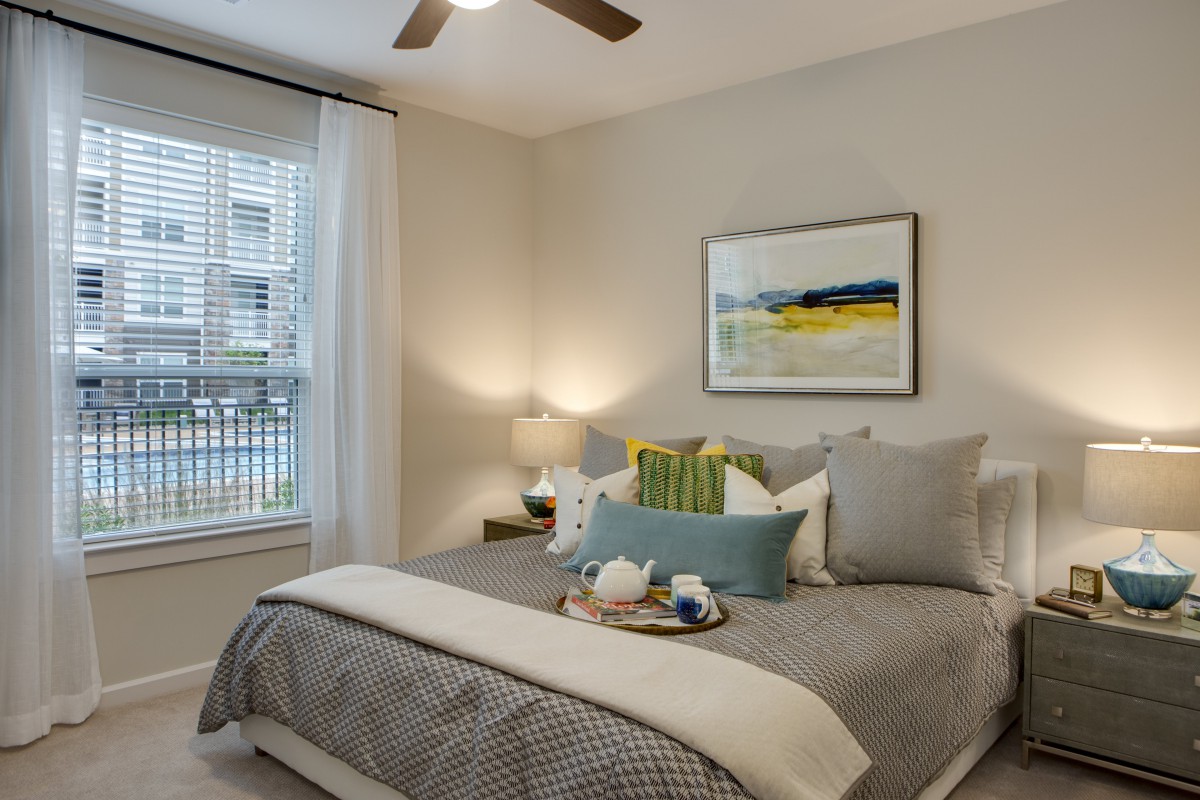 A cozy and well-decorated bedroom featuring a neatly made bed with pillows, artwork on the wall, and a bedside lamp next to a window.