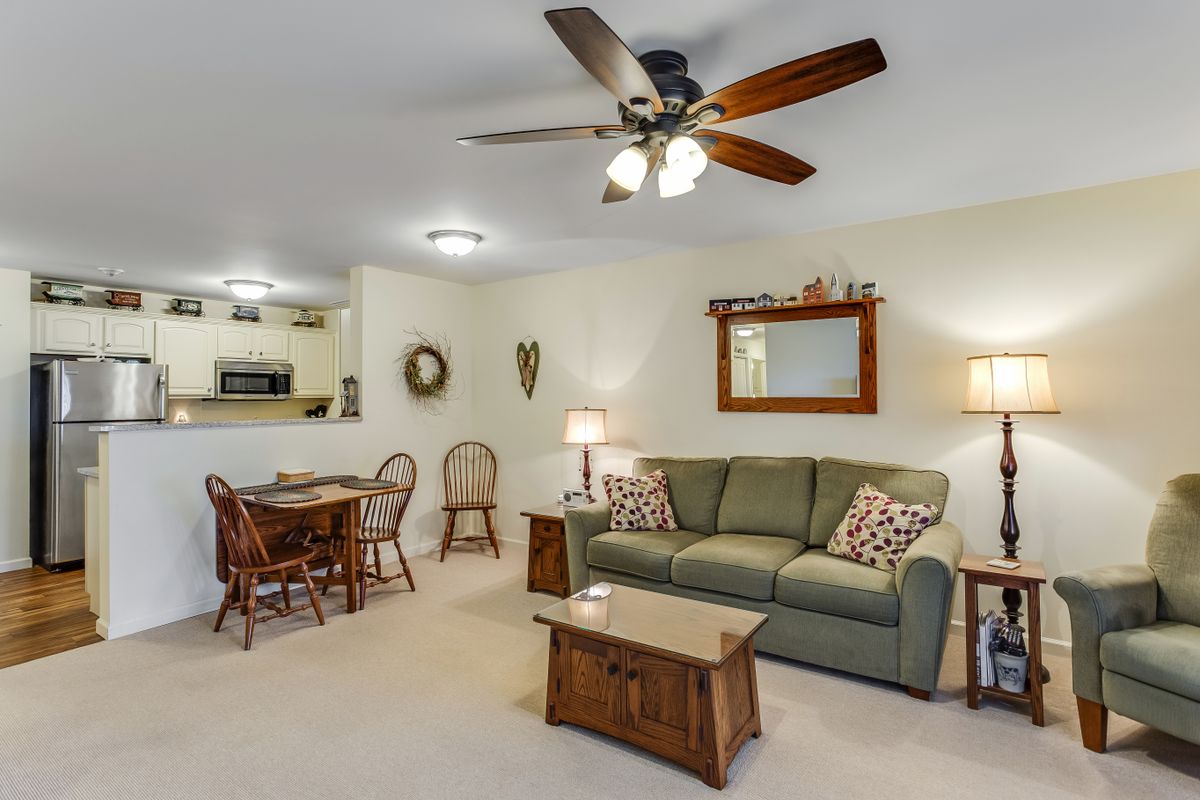 A cozy living room with a sofa, wooden coffee table, and dining area in the background, featuring a ceiling fan and warm lighting.