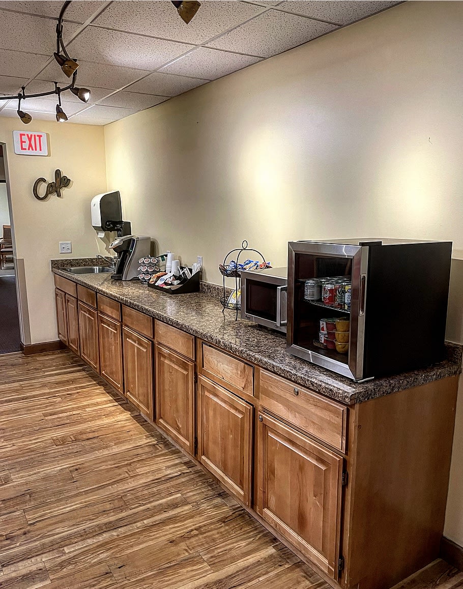 A kitchenette area with a long countertop, featuring a microwave, coffee maker, and snacks on wooden cabinets.
