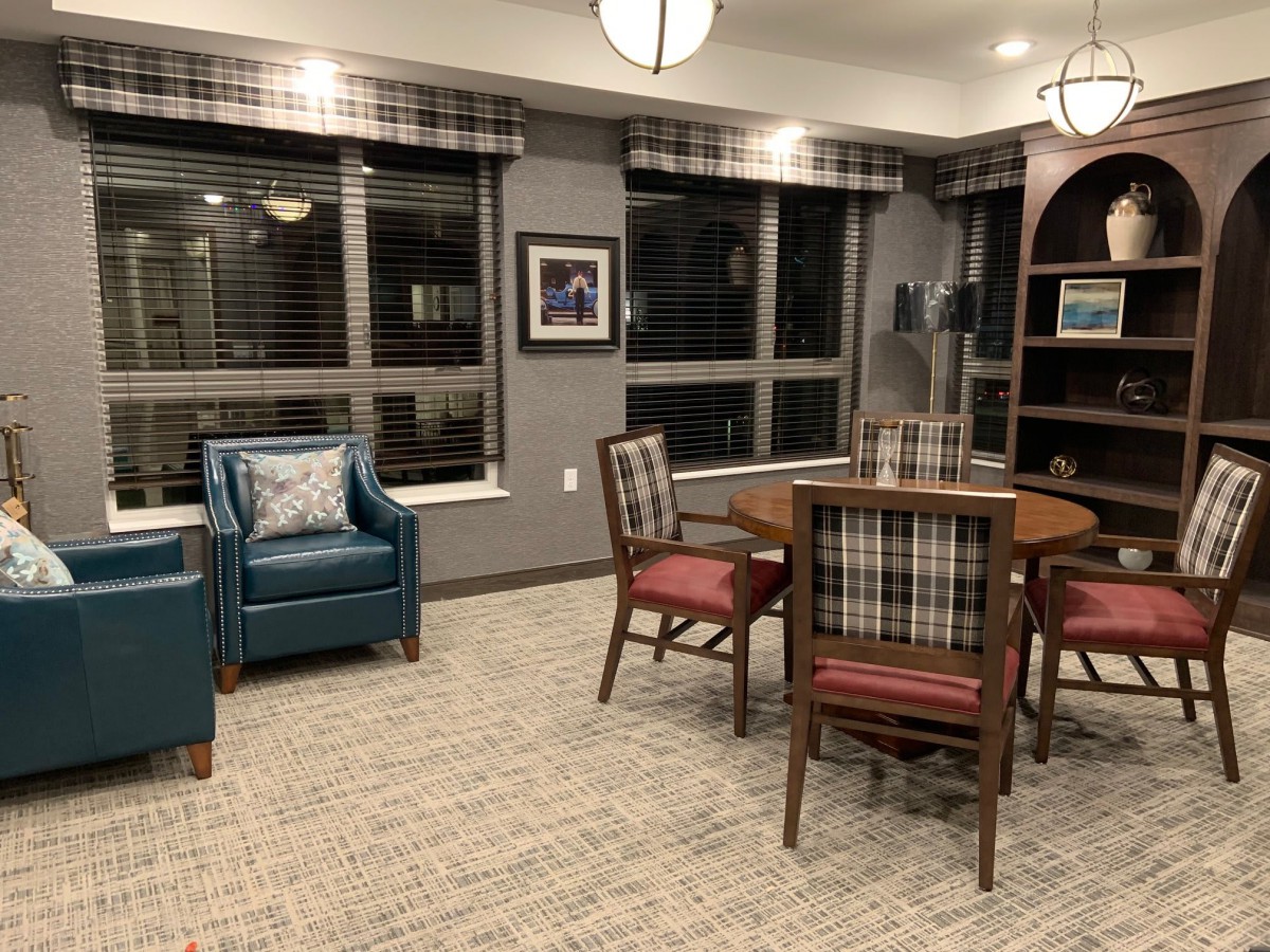 A cozy living room with a round table, armchair, and plaid-upholstered chairs under warm indoor lighting.