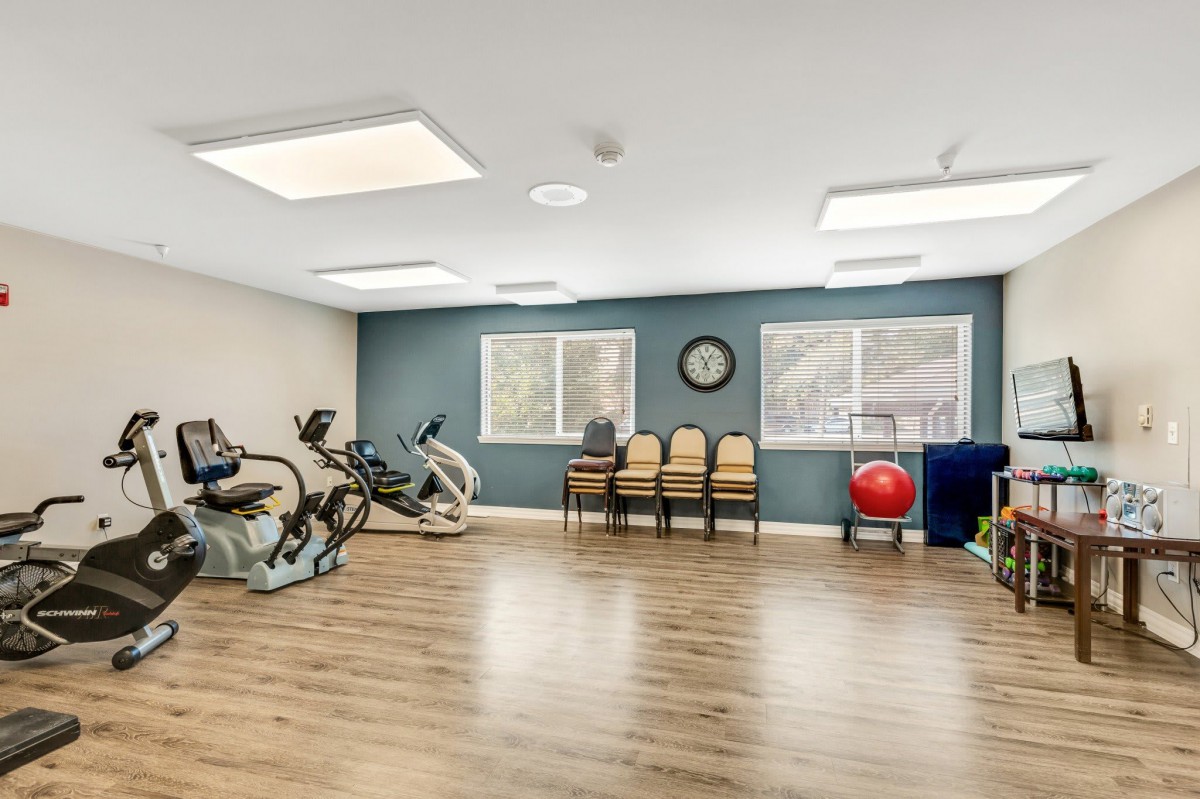 A spacious exercise room with gym equipment, including stationary bicycles and an exercise ball, featuring a wooden floor and windows for natural light.