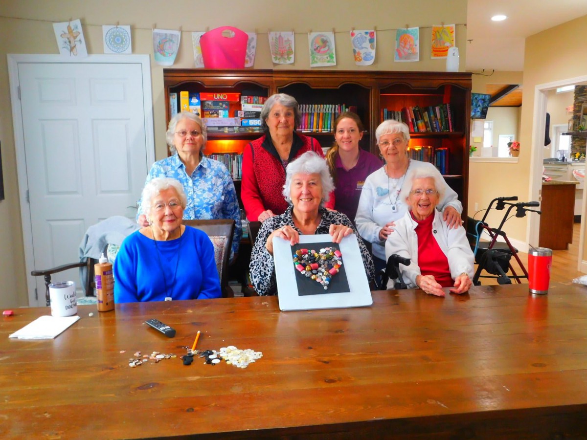 A group of elderly women smiling and posing with a crafted artwork in a cozy room.
