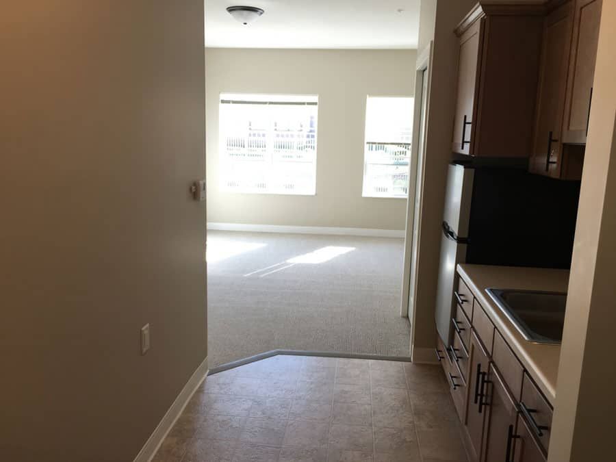View of a kitchen leading into a sunlit living room with large windows.