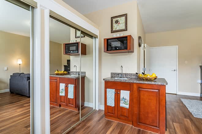A compact kitchenette with wood cabinets, a microwave, and a sink, featuring a mirrored closet door and a fruit bowl on the counter.