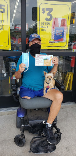A person wearing a mask sits outside with their small dog, holding a sign.