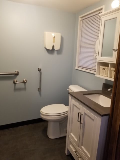 A clean and accessible bathroom with a sink, mirror, toilet, handrails, and a window.