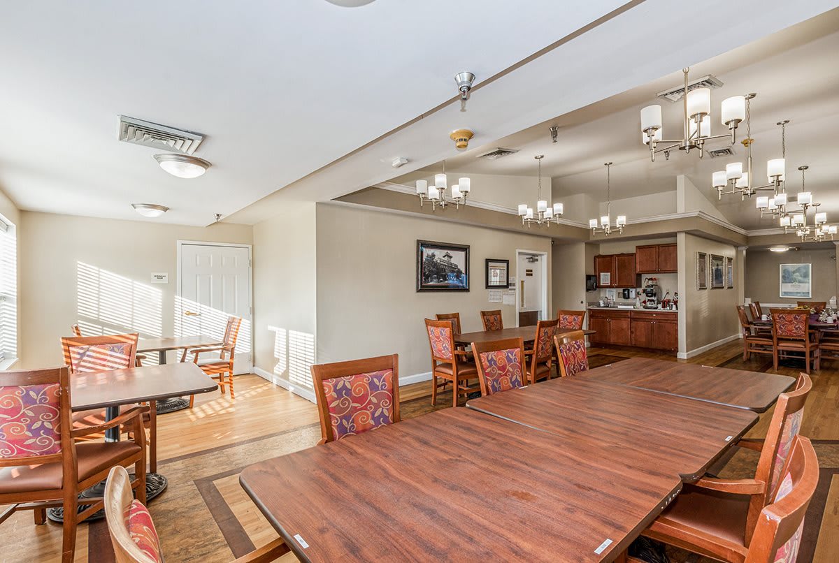 A spacious dining area with a large wooden table, chairs, and adjacent kitchen, well-lit with natural light and chandeliers.