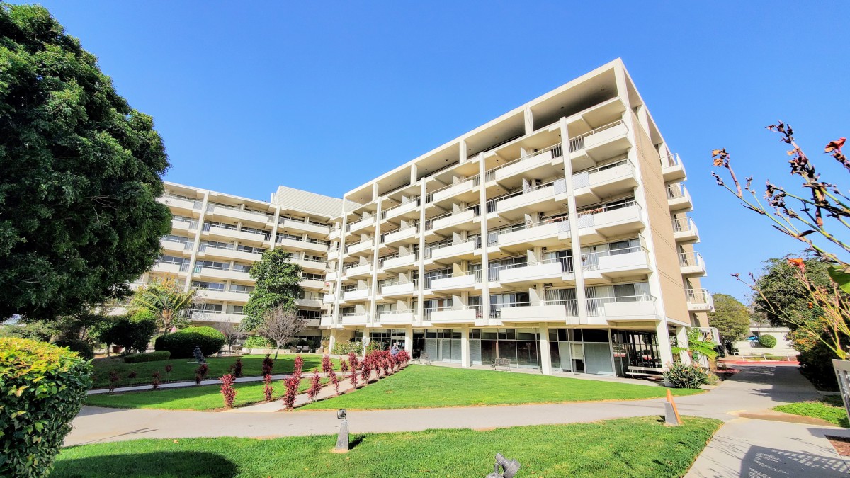 A modern apartment building with multiple balconies, surrounded by a well-maintained lawn under a clear blue sky.