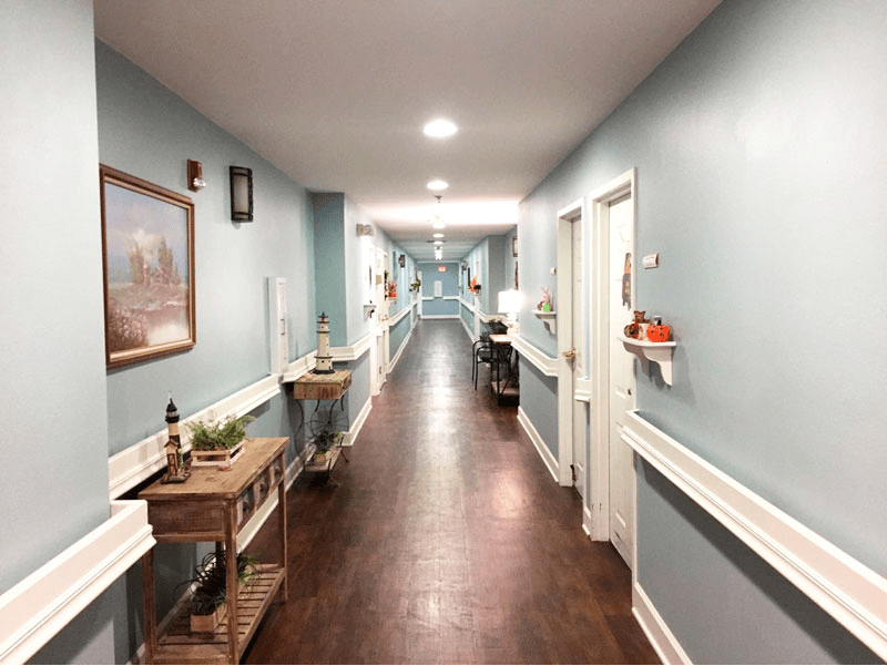 A well-lit nursing home hallway with decorated walls and a wooden floor.