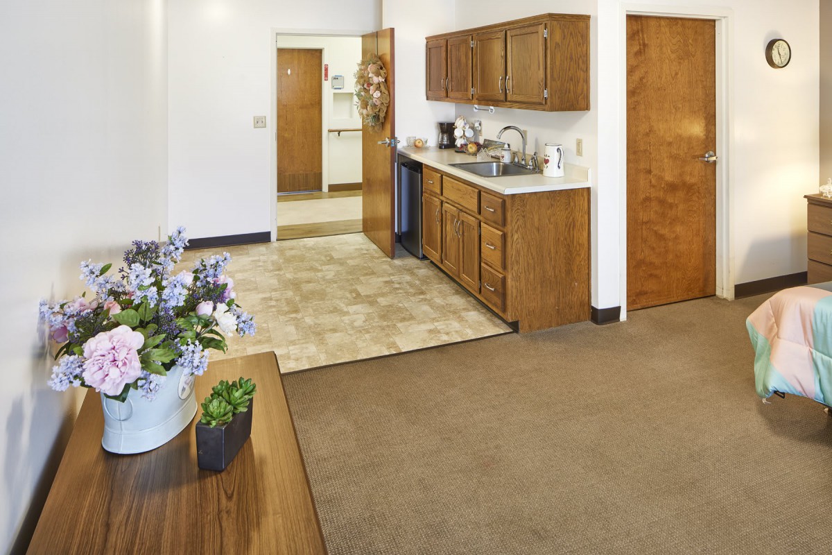 A clean apartment kitchen with wooden cabinets and a sink, featuring a small dining area adorned with potted flowers.