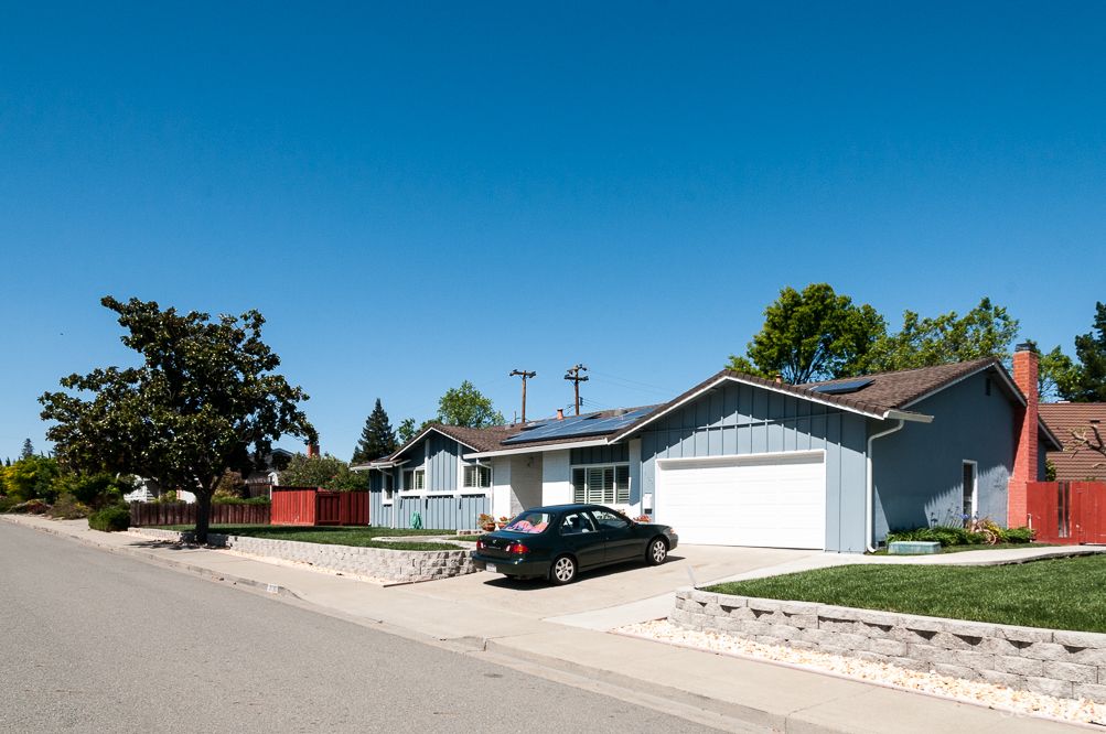 A suburban house with solar panels on the roof and a car parked in the driveway under a clear blue sky.