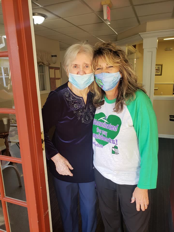 Two people wearing masks, standing side by side and smiling indoors.
