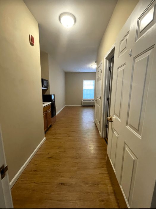 A long apartment hallway with wooden flooring, leading to a kitchen area and a window.
