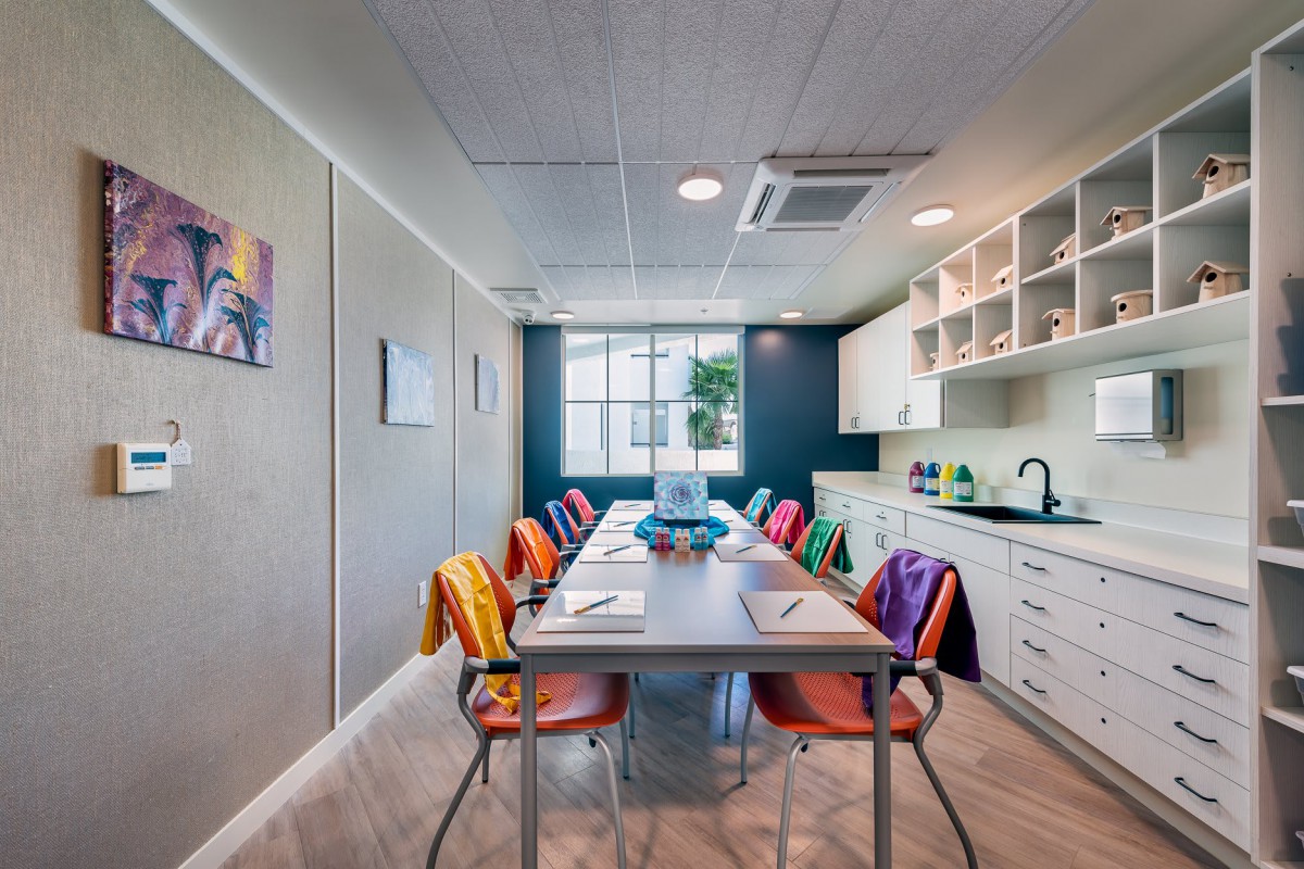 A modern meeting room with colorful chairs around a conference table, featuring a kitchenette and artwork on the walls.