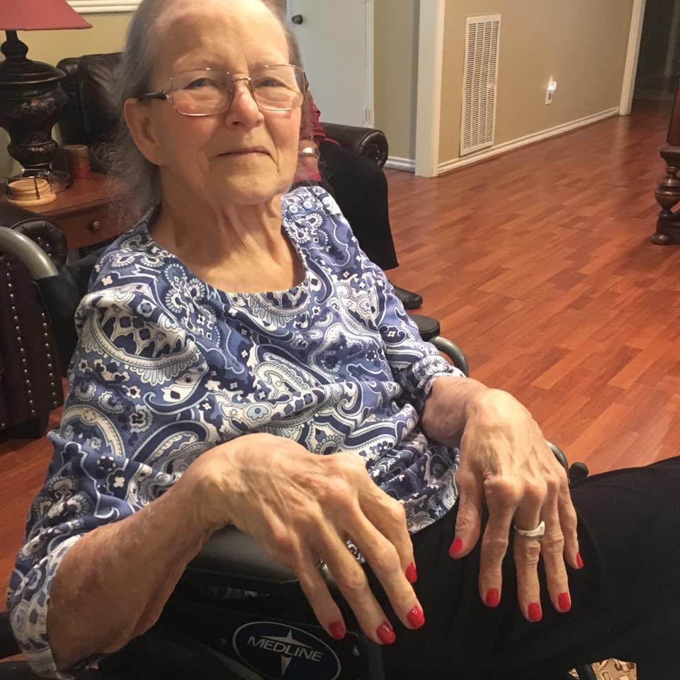 An older woman sitting in a wheelchair in a living room, wearing glasses and a patterned shirt.