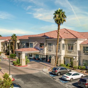 A sunny day view of a hotel building with palm trees and cars parked in the lot.