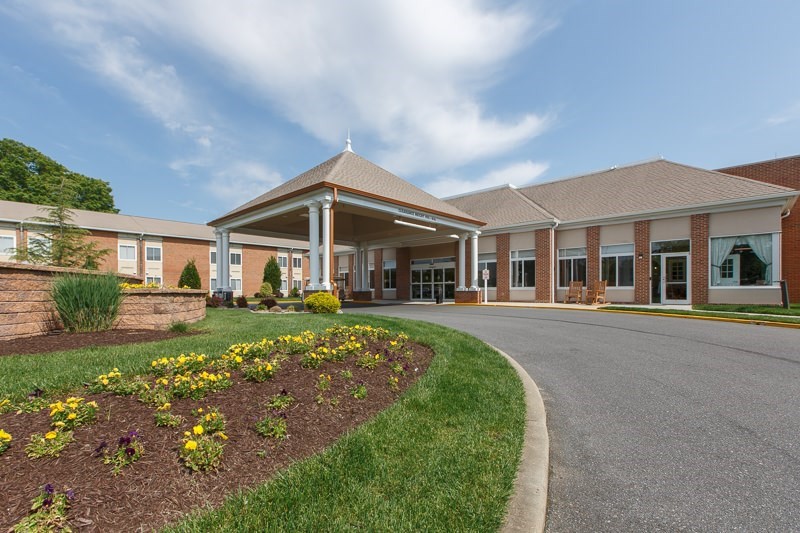 The image shows the entrance of a large building with a landscaped driveway and a partly cloudy sky.