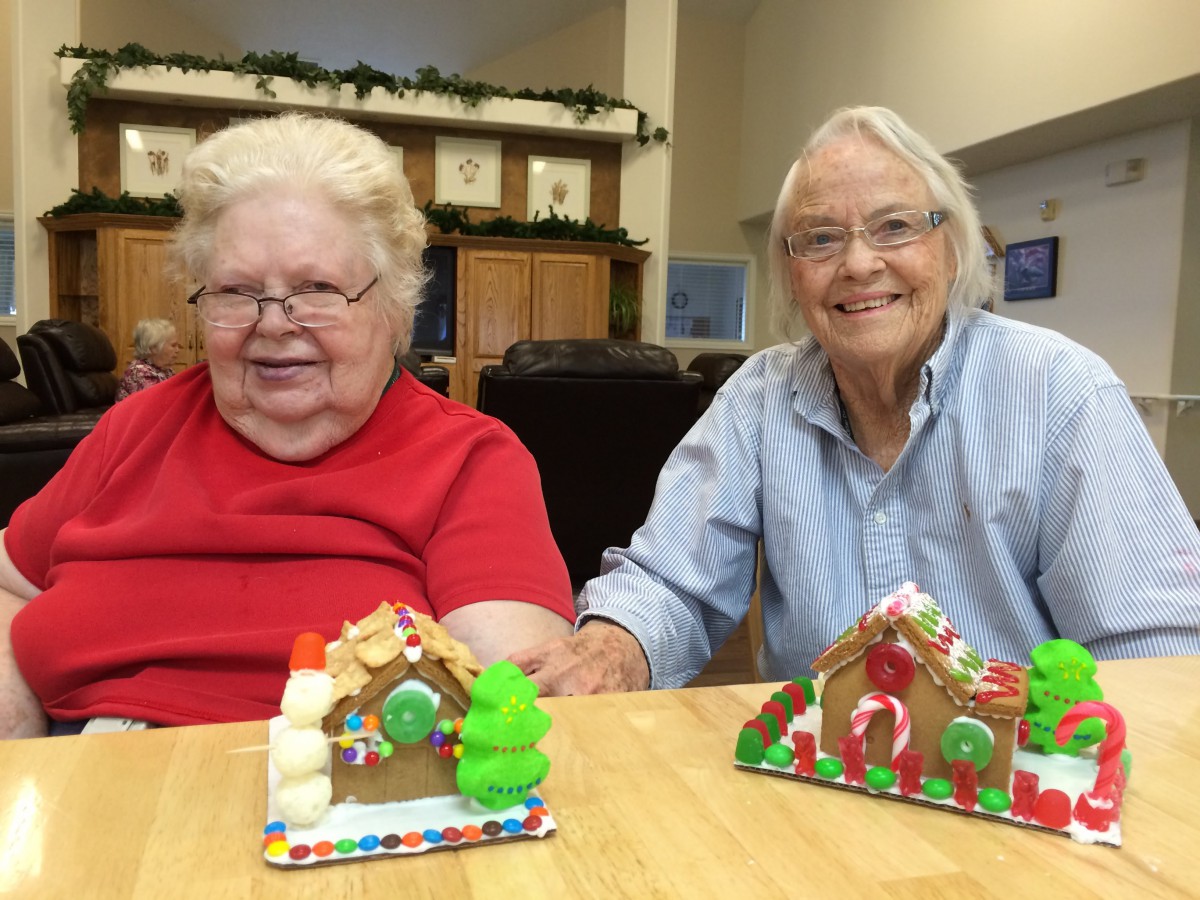 Two elderly women smiling while displaying their decorated gingerbread houses indoors.