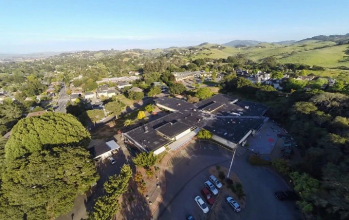 Aerial view of a complex of buildings surrounded by trees and hills with a parking lot in the foreground.